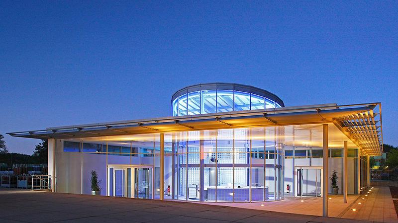 Neubau eines Empfangsgebaeudes Moderne Architektur eines Gebäudes bei Dämmerung. Die Fassade ist aus Glas und Metall, mit einem runden Dach. Sanfte Beleuchtung hebt die Struktur hervor und schafft eine einladende Atmosphäre. Um das Gebäude herum sind Pflanzen und eine gepflasterte Fläche sichtbar.