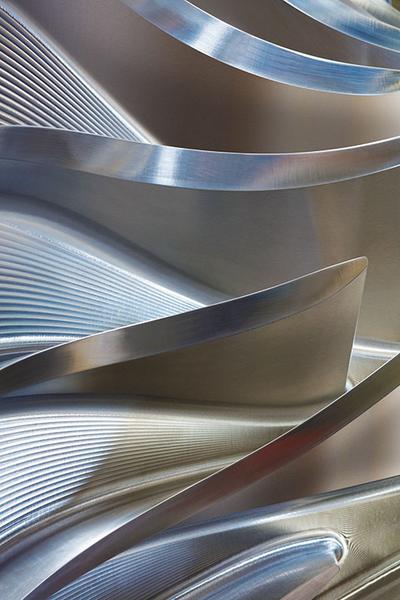 Turbinenlaufrad Eine Nahaufnahme von geschwungenen, polierten Metalloberflächen. Die Struktur zeigt wellenförmige und gebogene Elemente, die miteinander verbunden sind und verschiedene Lichtreflexionen erzeugen. Das Bild vermittelt eine dynamische und moderne Ästhetik durch die Kombination von Linien und Form.