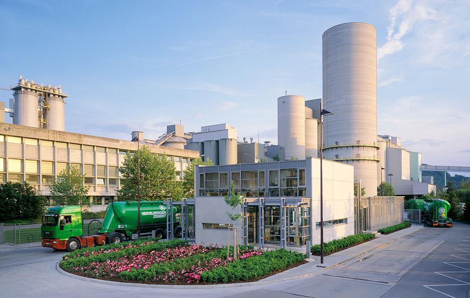 Werksansicht Ein modernes Industriegebäude mit großen Silos und einem Empfangsgebäude im Vordergrund. Grüne Lastwagen stehen vor dem Gebäude, umgeben von gepflegten Blumen und Bäumen. Der Himmel ist blau mit einigen Wolken.