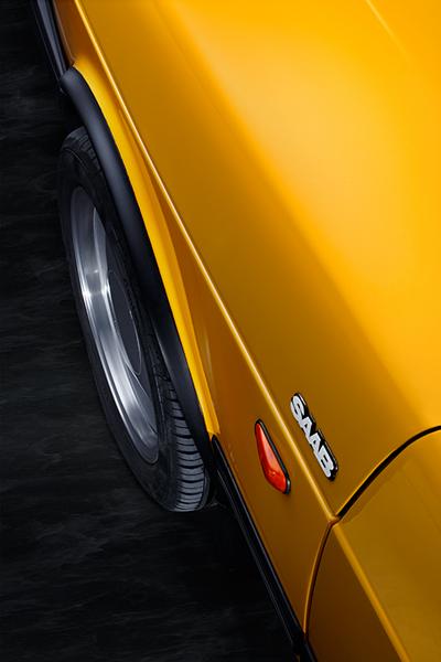Saab 900 i Cabriolet Kotflügel Ein gelber Saab-Auto mit einem glänzenden Lack. Die Aufnahme zeigt die Seitenansicht, wobei das Saab-Emblem und ein Rad sichtbar sind. Der Hintergrund ist dunkel und hebt die Farbe des Fahrzeugs hervor.