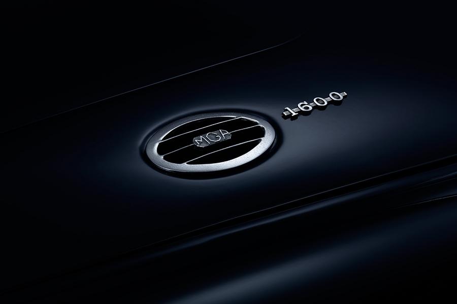 MGA 1600 Roadster Hutze Ein schimmerndes Logo und die Modellbezeichnung "MGA 1600" auf einer dunklen Fahrzeugoberfläche. Das Logo hat eine runde Form mit horizontalen Streifen und ist in silberner Farbe gehalten.