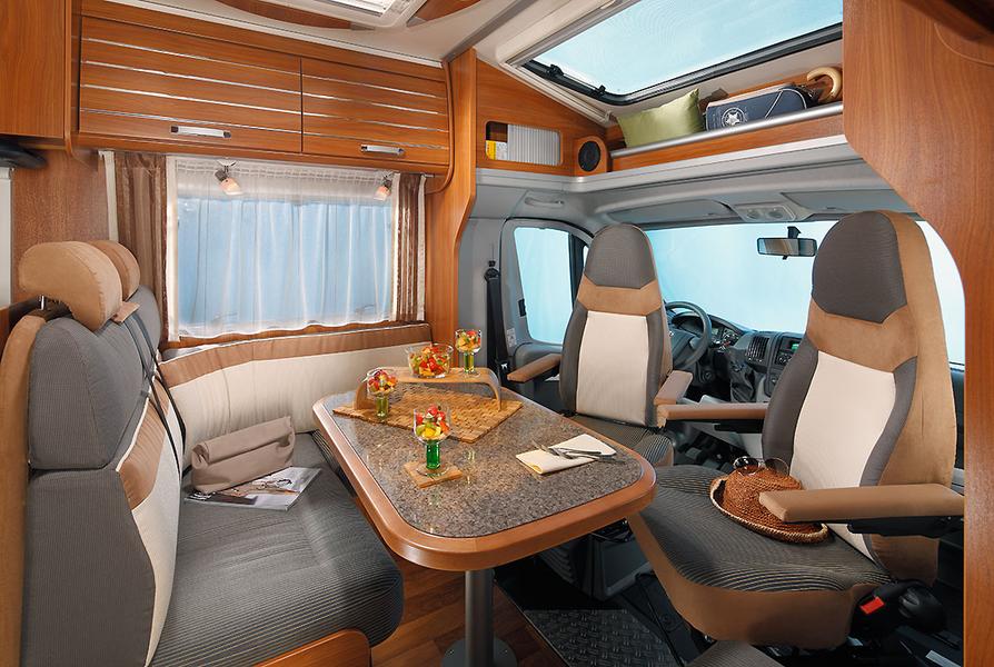 Wohnmobil Fahrerhaus Innenansicht eines Wohnmobils mit einem Esstisch, umgeben von zwei Sitzgruppen. Auf dem Tisch stehen Snacks und Getränke. Die Wände sind mit Holzverkleidung ausgestattet, und große Fenster sorgen für genügend Licht. Im Hintergrund ist der Fahrersitz zu sehen.
