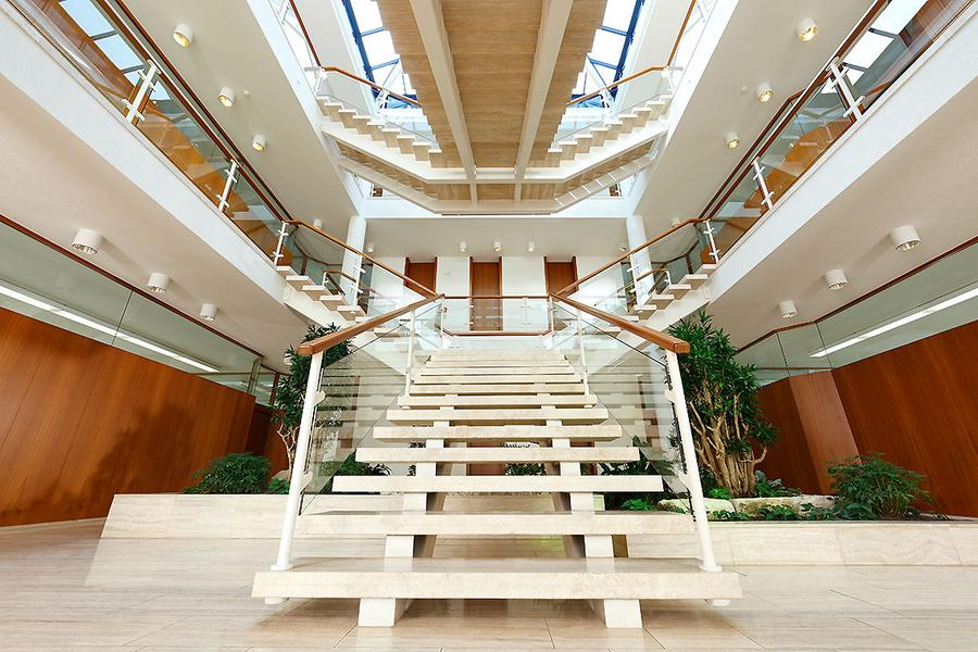 Treppenaufgang Eine moderne Treppe aus Holz und Glas führt in einen hellen Innenraum mit mehreren Etagen. Die Wände sind warmbraun, und es gibt Pflanzen im Raum. Oberlichter lassen natürliches Licht herein.