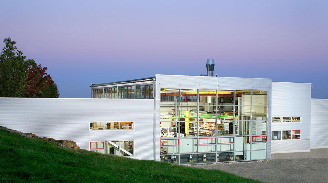Werksansicht Moderne Architektur eines Gebäudes mit großen Glasfenstern. Die Fassade ist weiß, umgeben von Gras und Bäumen im Hintergrund. Der Himmel ist bei Dämmerung in sanften Farben gehalten.