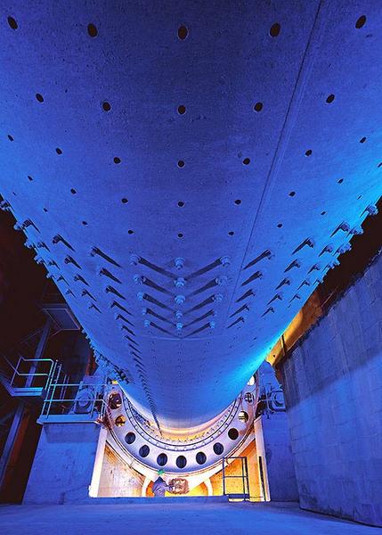 Zementmuehle Blick auf die Unterseite eines Tunnelbohrers, beleuchtet in blauen und gelben Tönen. Die Struktur zeigt runde Löcher und mechanische Komponenten, die in einem unterirdischen Raum positioniert sind. Teile der Tunnelbau-Infrastruktur sind im Hintergrund sichtbar.