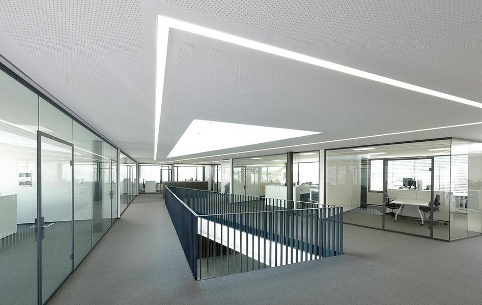 Schmieder Gebaeude Moderne Bürolandschaft mit Glaswänden und einem offenen Bereich. Helle, minimalistische Gestaltung mit natürlichem Licht, das durch ein großes Dachfenster strömt. Ein erhöhter Gang führt zwischen den Büroräumen. Der Boden ist mit grauem Teppich ausgelegt.