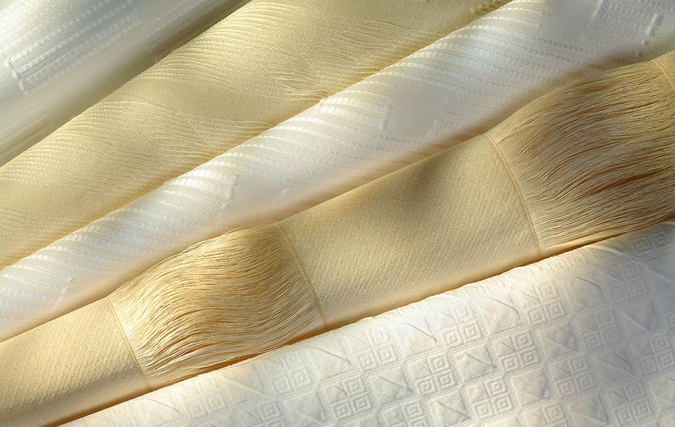 Vier Lagen von Stoffen in verschiedenen cremigen Farbtönen, die sanft übereinander liegen. Die Texturen variieren, mit glatten und strukturierenden Mustern, wodurch ein elegantes und leicht schimmerndes Erscheinungsbild entsteht.