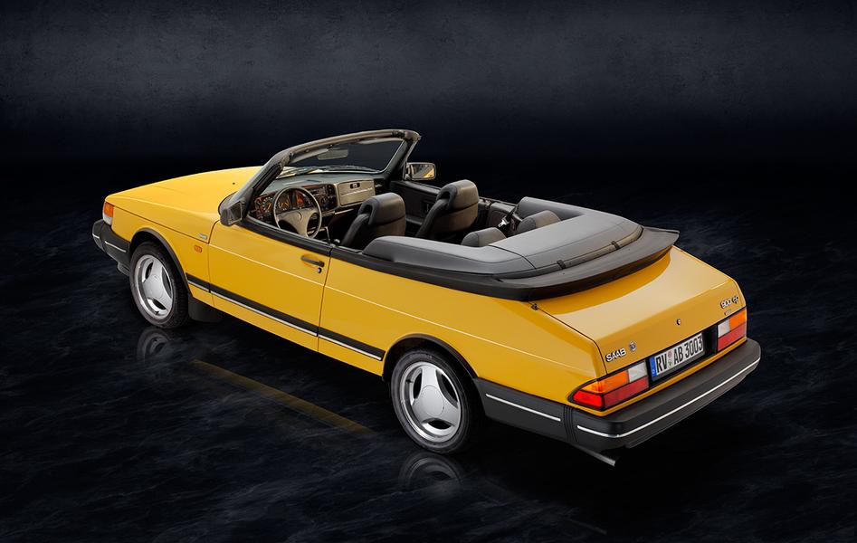 Saab 900 i Cabriolet Heck Ein gelbes Cabriolet mit schwarzem Verdeck steht vor einem dunklen Hintergrund. Die Rückansicht zeigt die charakteristischen Rundungen des Autos und die Designs der Felgen. Innenraumdetails sind teilweise sichtbar.