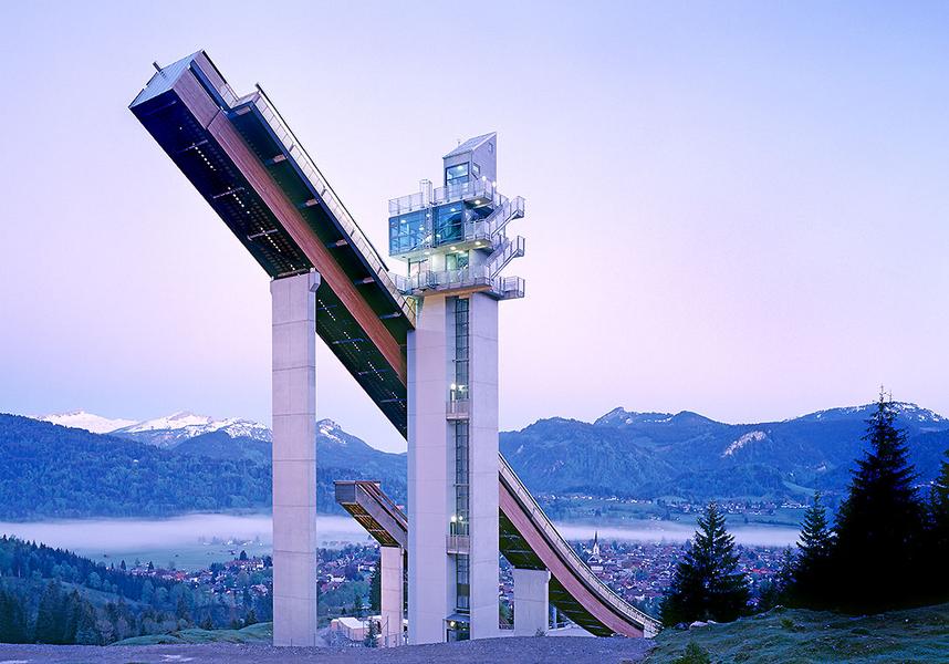 Audi Arena Oberstdorf Eine Skisprungschanze steht majestätisch vor einem sanften Bergpanorama. Der hellblaue Himmel im Hintergrund verdeutlicht die Morgenstunde, während die umliegenden Wälder und das Tal leicht in Nebel gehüllt sind.