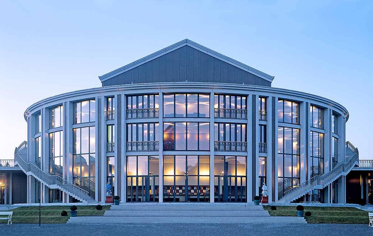 Festspielhaus Fuessen Eine runde, moderne Gebäudeansicht mit großen Glasfenstern. Zwei Treppen führen zum Haupteingang. Der Himmel ist hellblau und der Garten vor dem Gebäude ist ordentlich gestaltet. Die Architektur kombiniert traditionelle und zeitgenössische Elemente.