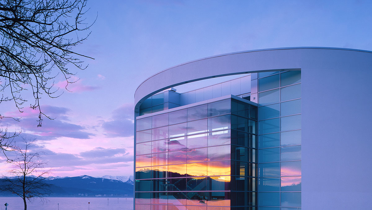 Spielbank Lindau Moderne Gebäude mit großen Glasfenstern, reflektierend die Farben eines Sonnenuntergangs. Im Hintergrund sind Berge und ein ruhiger See sichtbar. Die Szene vermittelt eine friedliche Atmosphäre.