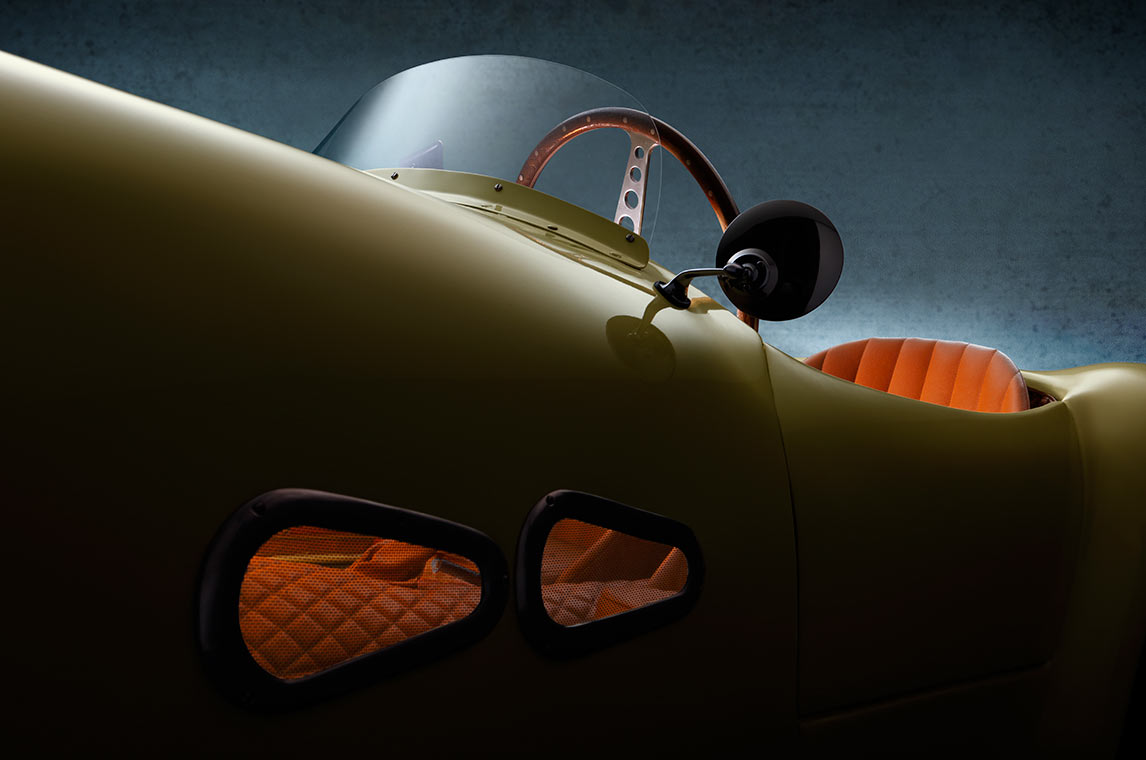 FIAT 500 Graziani MM Kotfluegel Nahaufnahme eines klassischen Autos mit einer geschwungenen Form. Die Karosserie ist in einem gelben Farbton, und die Sitze sind in einem auffälligen Orangeton gehalten. Der Innenraum zeigt ein modernes Lenkrad und eine Windschutzscheibe, während die Hintergrundfarbe dunkel und strukturiert ist.
