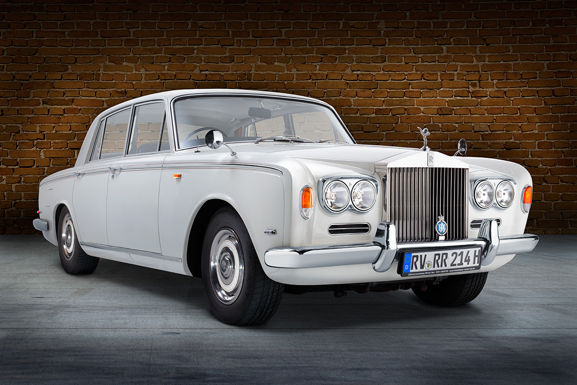 Rolls Royce Silver Shadow Front Ein klassisches weißes Auto, ein Rolls-Royce, steht vor einer Backsteinmauer. Die Front weist markante Scheinwerfer und das bekannte Kühleremblem auf. Das Fahrzeug hat eine elegante und luxuriöse Erscheinung.