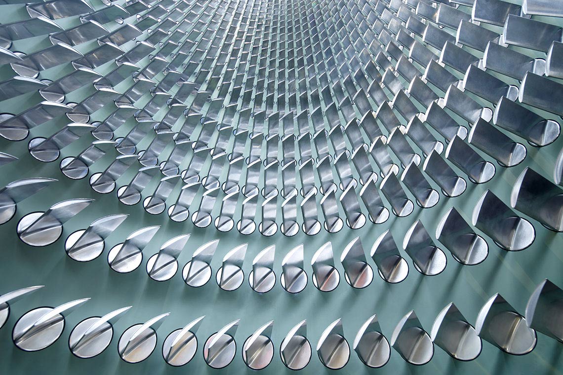Schaufeltraeger Eine abstrakte Struktur mit sich spiralförmig anordnenden, glänzenden Metallteilen. Die Elemente sind halbrund und reflektieren das Licht, was einen dynamischen Eindruck erzeugt. Der Blickwinkel ist von unten nach oben gerichtet, wodurch die Tiefe der Form betont wird.