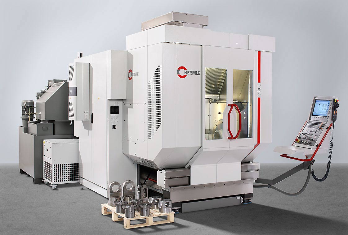 CNC Bearbeitungsanlage Eine moderne CNC-Maschine in einem industriellen Umfeld. Die Maschine hat eine große, weiße Gehäusekonstruktion mit Fenster, einem Bedienfeld und einem Stapel von bearbeiteten Metallteilen davor. Im Hintergrund ist eine weitere Maschine sichtbar.