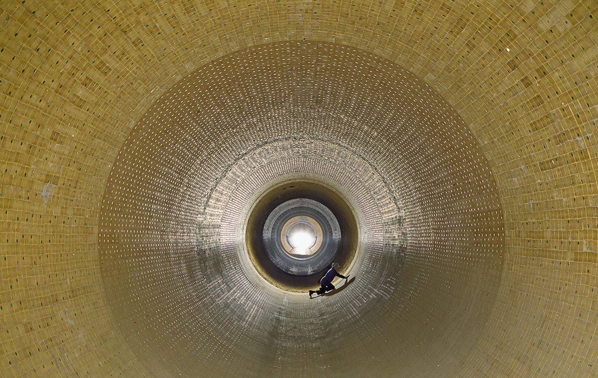 Drehofenausmauerung Eine perspektivische Ansicht des Innenraums eines großen, zylindrischen Behälters aus Steinen oder Platten. In der Mitte ist eine Lichtquelle sichtbar, während eine Person am Boden arbeitet. Die Wände sind rhythmisch strukturiert und reflektieren das künstliche Licht.