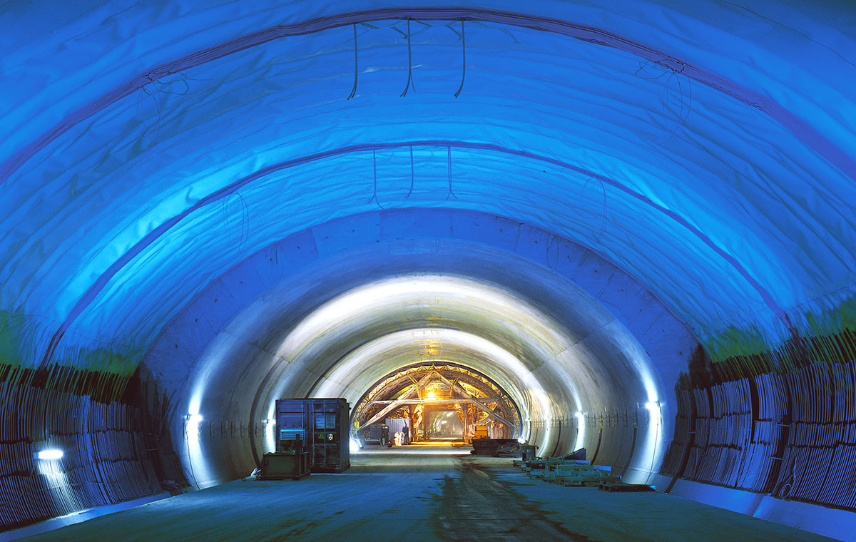 Autobahntunnel A81 im Bau Ein beleuchteter Tunnel mit blauen Wänden und einem gewölbten Dach. Im Hintergrund sind einige Baugeräte und Personen zu sehen. Der Boden ist eben und der Tunnel scheint gut strukturiert zu sein.