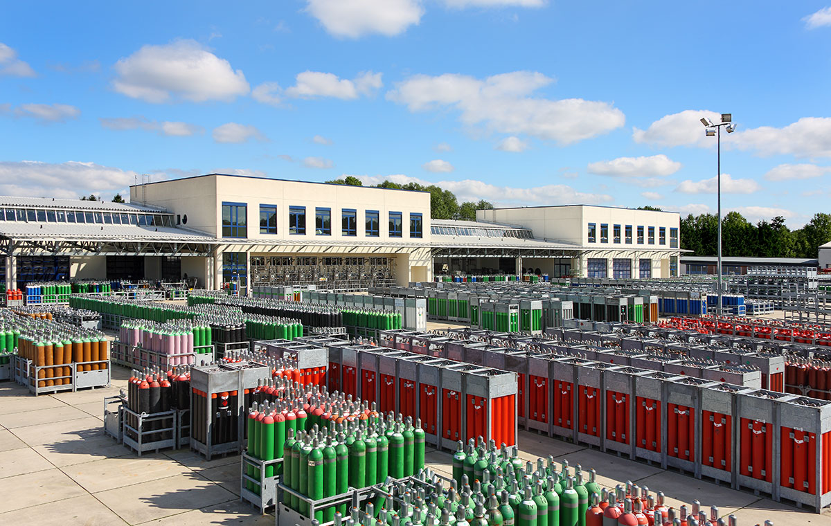 Werksansicht Eine weitläufige Lagerfläche mit zahlreichen Gasflaschen in unterschiedlichen Farben. Im Hintergrund sind moderne Gebäude und ein blauer Himmel mit wenigen Wolken zu sehen.