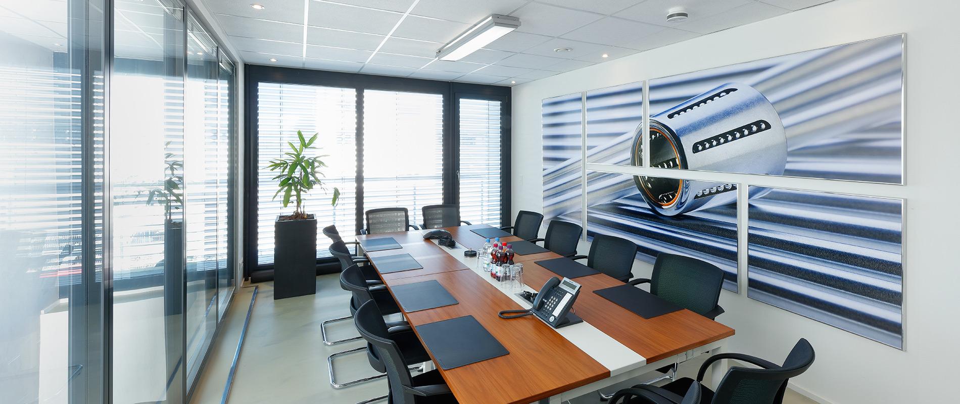Grossformatdruck Ein modern gestalteter Konferenzraum mit einem langen Tisch, umgeben von Stühlen. An der Wand hängt ein großes Bild eines Flugzeugmotors. Große Fenster lassen viel Tageslicht herein, und in einer Ecke steht eine Pflanze.