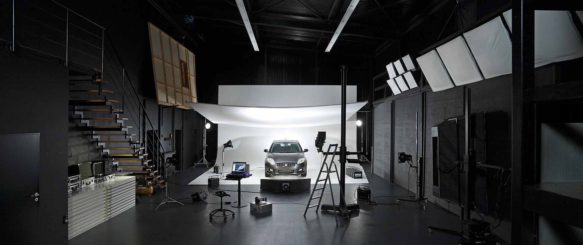 Firmengebaeude Becker Studios Ein modernes Fotostudio mit einem abgestellten Auto im Hintergrund. Die Szenerie ist gut beleuchtet, mit mehreren Lampen und einem weißen Hintergrund, der das Fahrzeug ins Licht rückt. An der Seite sind verschiedene Kamera- und Studioausrüstungen sichtbar.