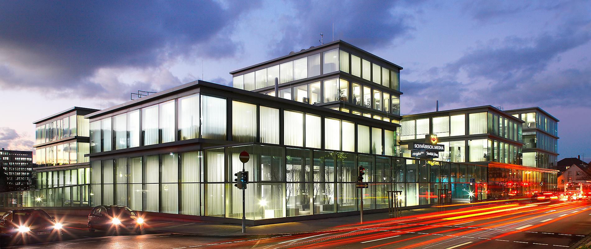 Moderne Architektur eines Glasgebäudes bei Dämmerung, umgeben von Straßenlichtern und Fahrzeugen. Das Gebäude hat viele Fenster und ist stimmungsvoll beleuchtet.
