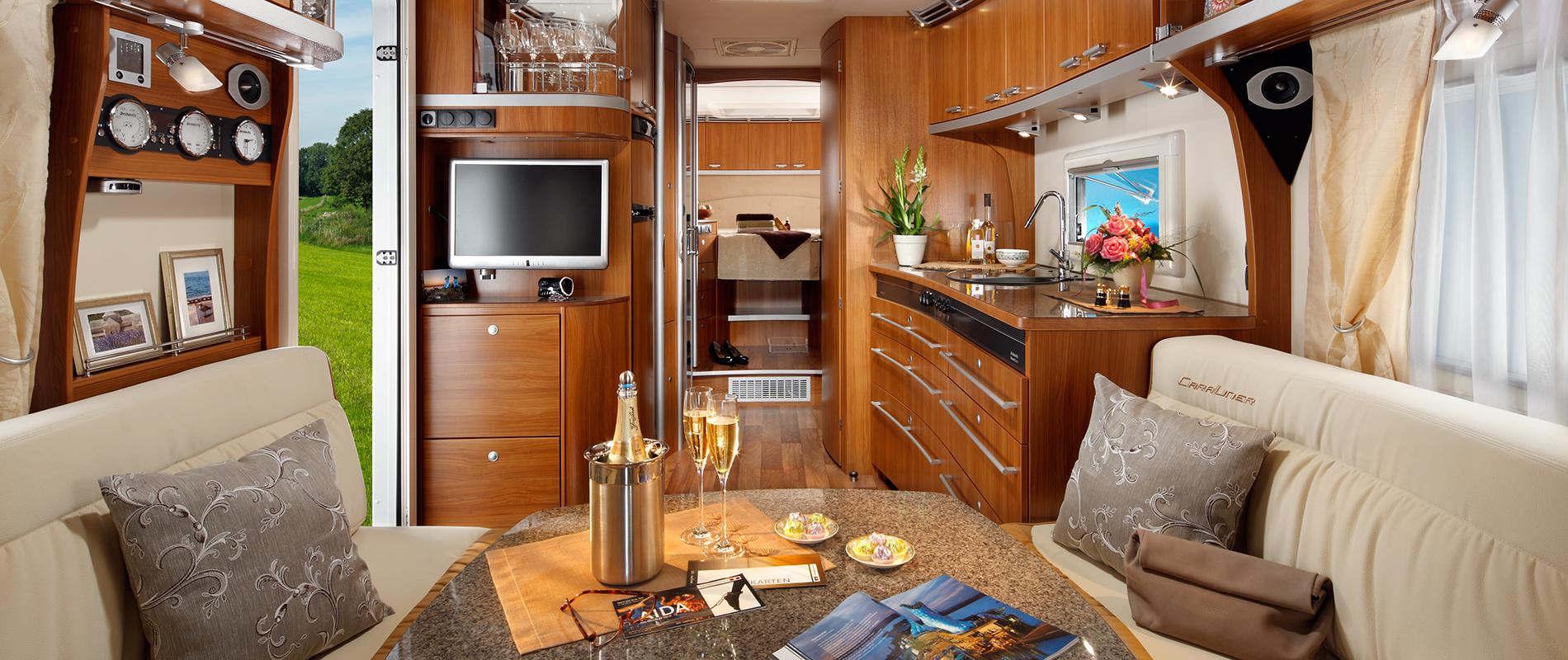 Innenansicht eines luxuriösen Wohnmobils mit Holzmöbeln. Ein Essbereich mit Gläsern und Snacks ist sichtbar, ebenso eine Küche und ein Wohnbereich. Ein Fernseher hängt an der Wand, und große Fenster sorgen für viel Licht. Draußen ist ein grünes Feld zu sehen.
