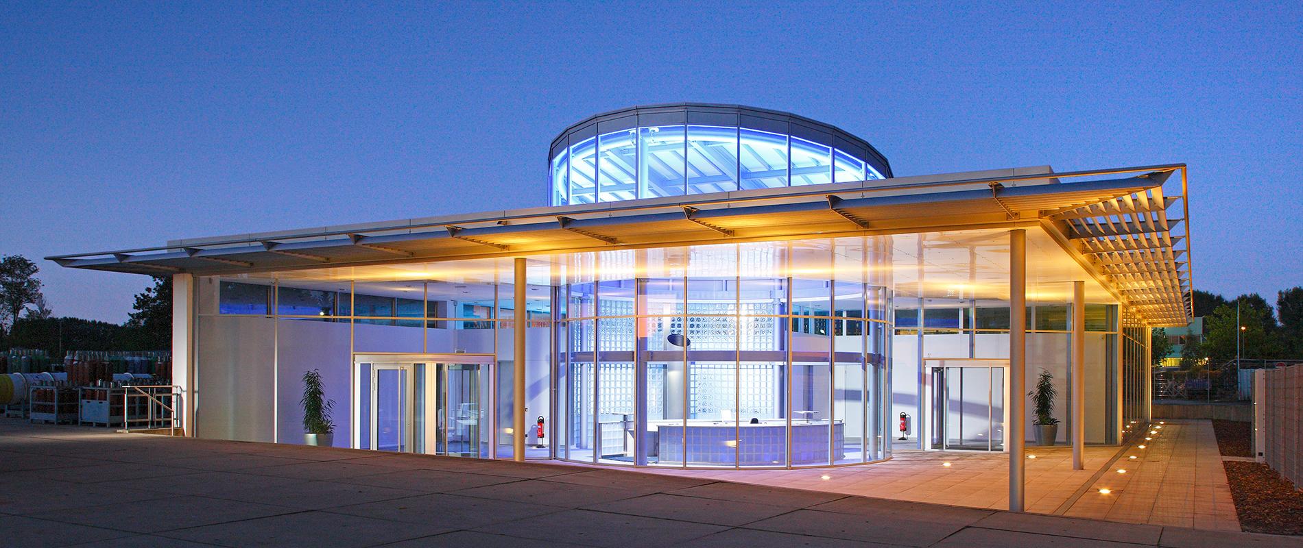 Moderne Gebäude mit gläserner Fassade und runder Kuppel im oberen Bereich. Die Fassade ist beleuchtet und reflektiert leicht das Licht des Abendhimmels. Vor dem Gebäude ist ein großzügiger Eingangsbereich zu sehen.