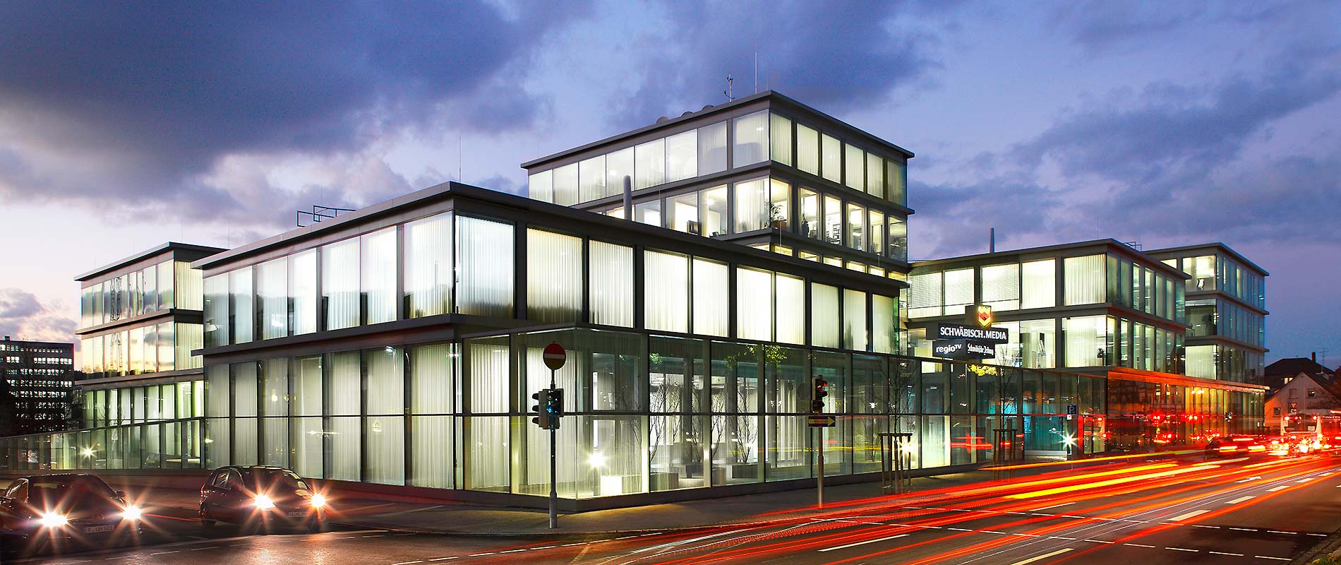 Redaktionsgebaeude Moderne Bürogebäude mit Glasfassade bei Dämmerung. Die Architektur ist klar und geometrisch, umgeben von Straßen mit leuchtenden Fahrzeuglichtern. Der Himmel ist bewölkt und düster, was die beleuchteten Fenster hervorhebt.