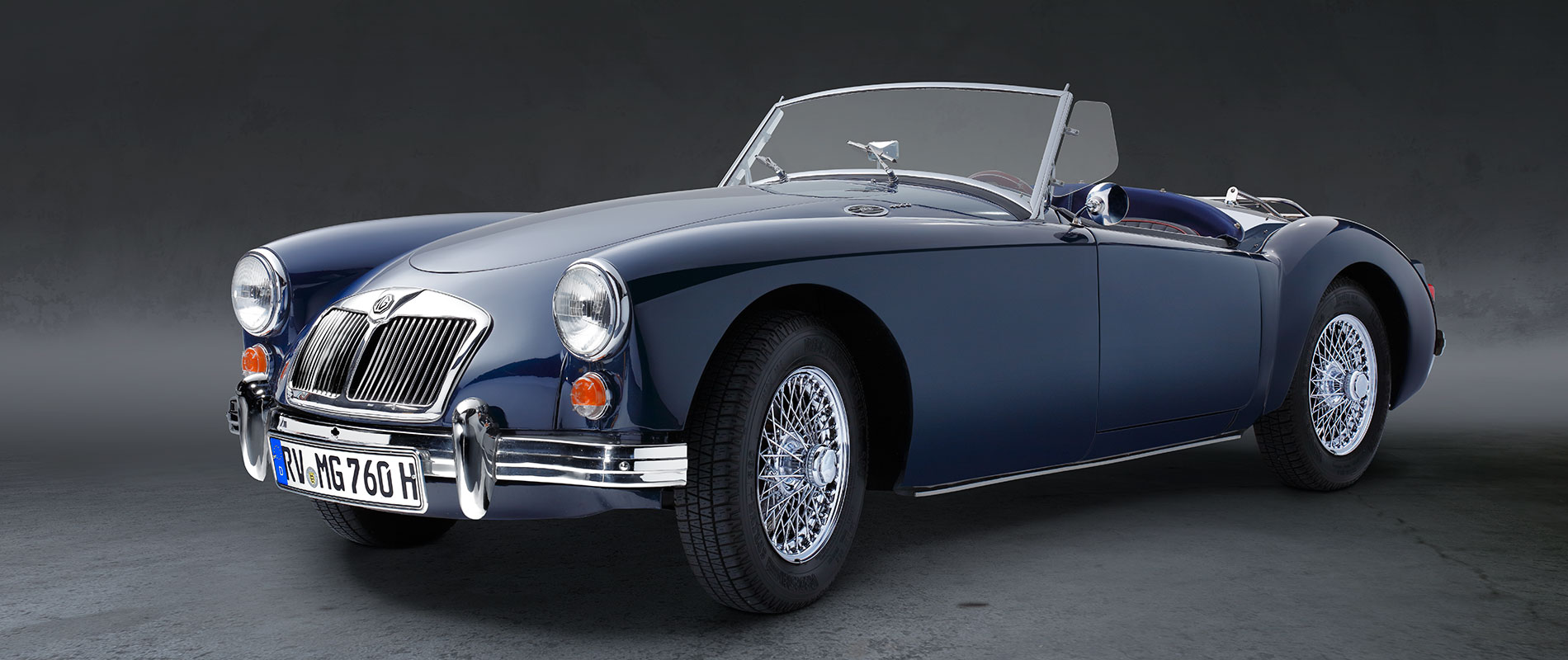 MAG Front Ein blauer Sportwagen im Cabriolet-Stil steht auf einer neutralen Fläche. Das Auto hat einen klassischen Look mit verchromten Details und einem offenen Verdeck. Die Felgen sind verchromt und auffällig gestaltet.