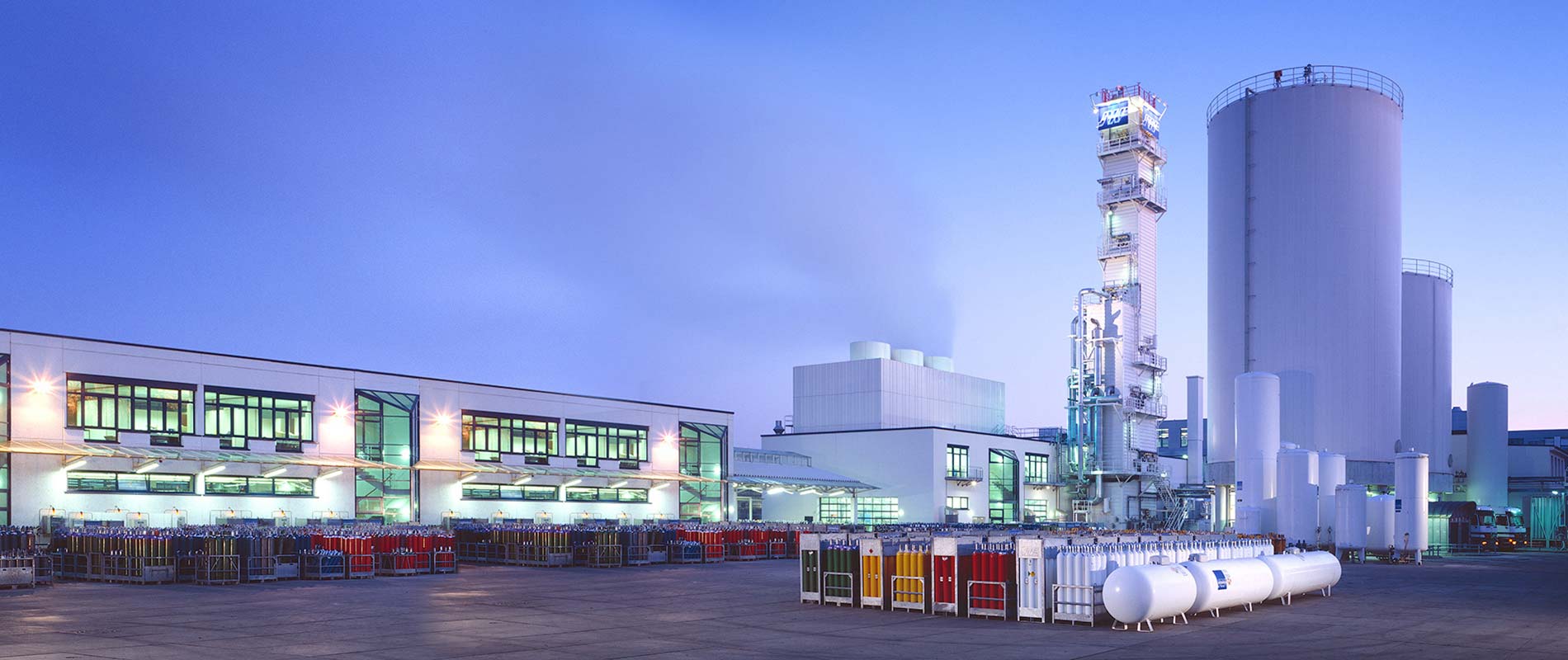 Werksansicht Industriegelände bei Dämmerung mit großen Lagertanks, Produktionsgebäuden und Gaszylindern auf dem Boden. Die Gebäude sind modern und gut beleuchtet.