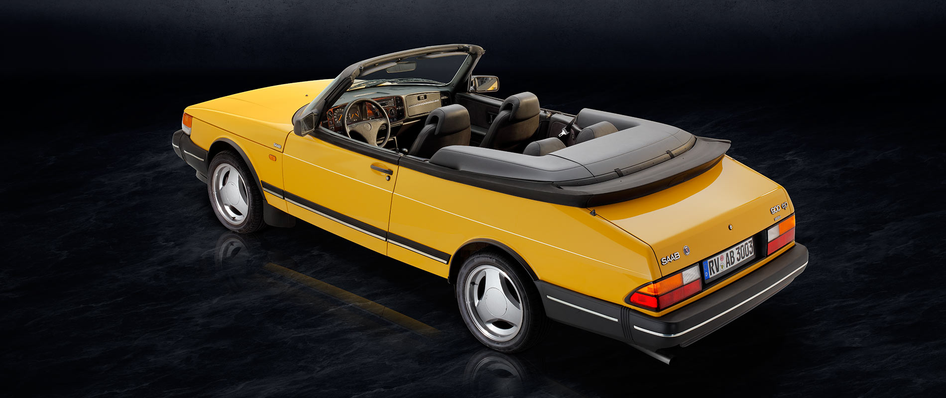 Saab 900 i Cabriolet Heck Ein gelbes Cabrio mit schwarzem Innenraum und gefaltetem Verdeck steht auf einer dunklen Oberfläche. Die Rückansicht zeigt die charakteristischen des Autos, einschließlich der Felgen und der Heckleuchten.